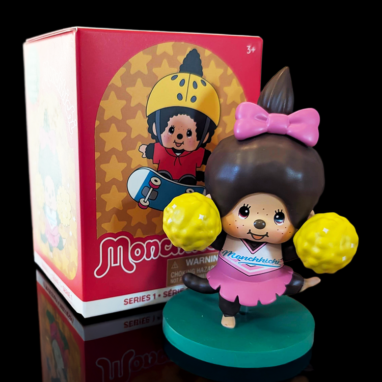 Sekiguchi Girl Monchhichi Cheerleader vinyl figure in pink outfit with yellow pom-poms on green stand next to box