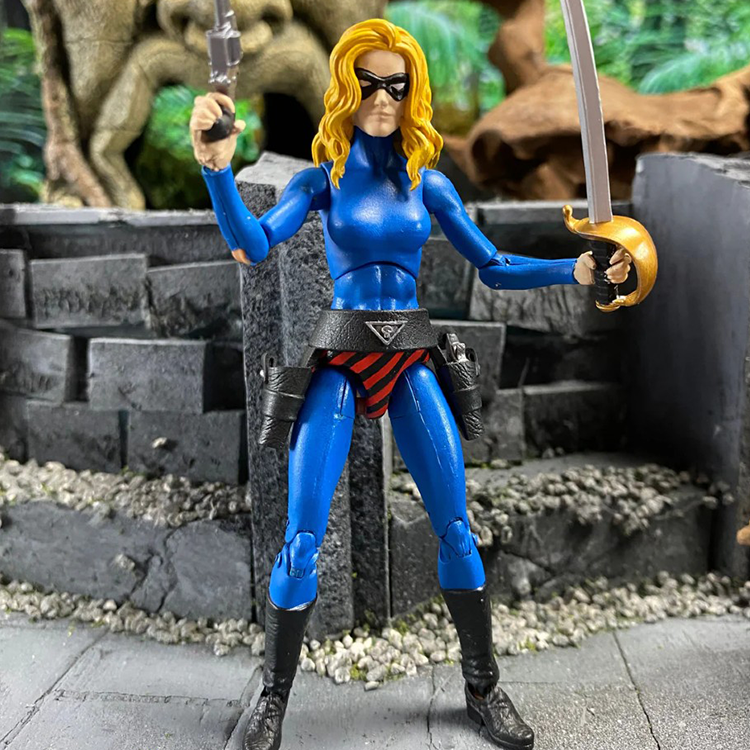 Articulated female action figure in blue suit holding a sword and pistol, standing on stone ground with greenery backdrop.