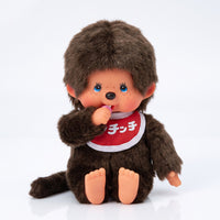 Brown Monchhichi doll with blue eyes red bib and one hand raised to its mouth