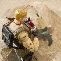 Blond military action figure holding a red-labeled device beside a rifle on sandy ground