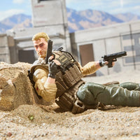 Blond soldier action figure lying in sand with a rifle and pistol near desert ruins