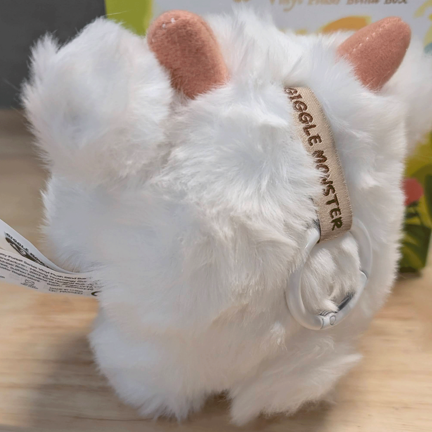 OHKU Giggle Monster plush with soft white fur, pink horns, and fabric tag, shown from rear on wooden surface