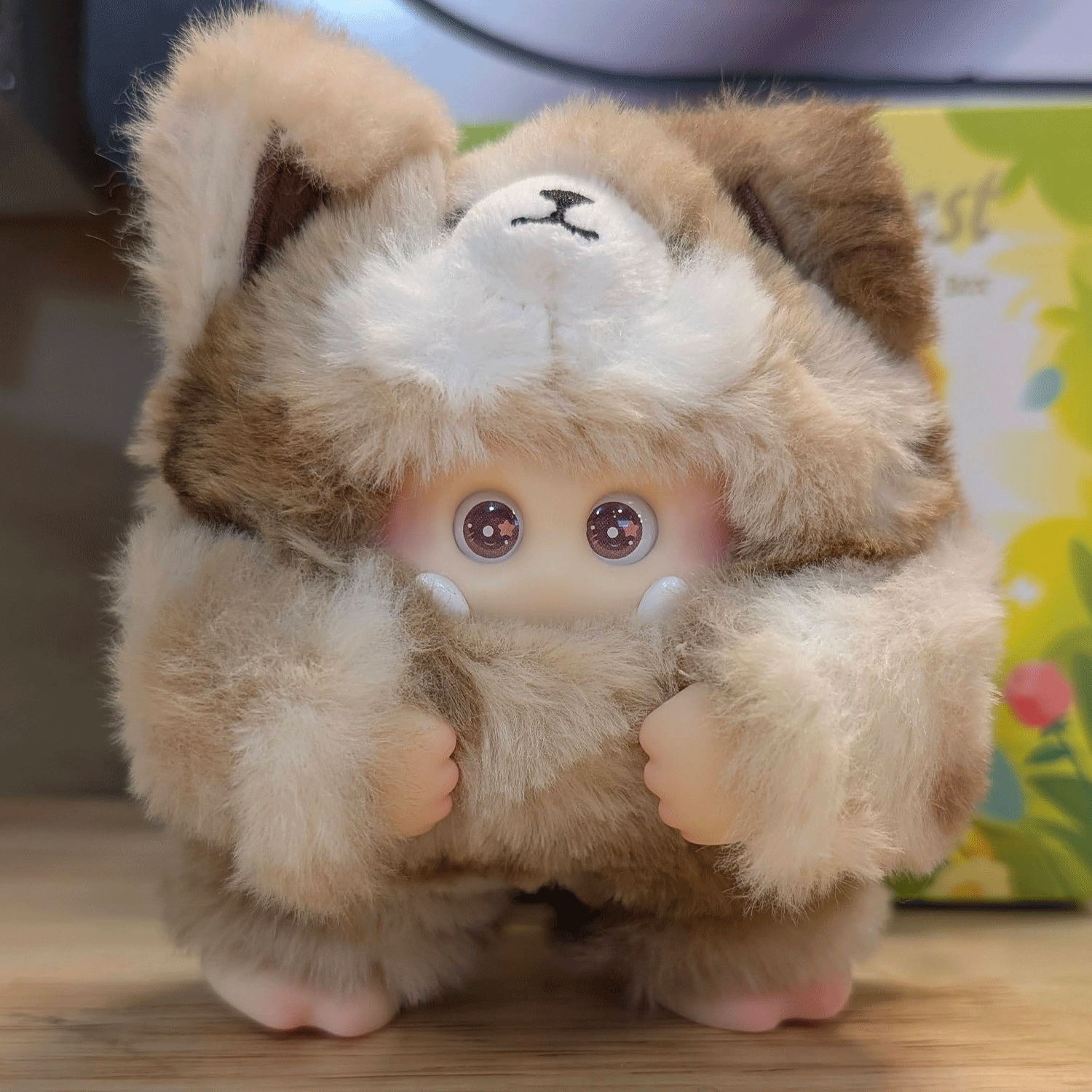 OHKU Giggle Monster vinyl plush with wide eyes in fluffy tan and cream furry creature outfit on wooden surface