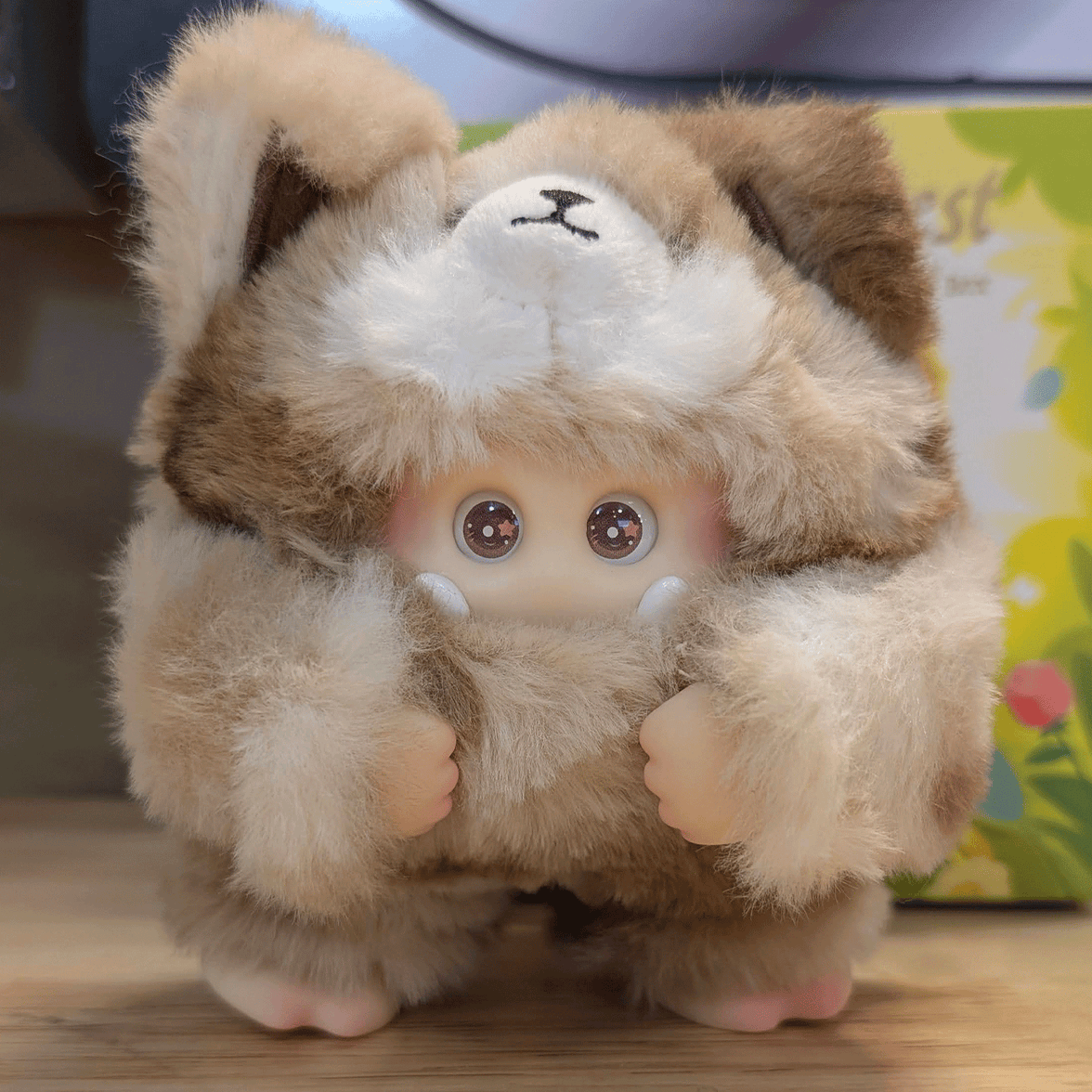 OHKU Giggle Monster vinyl plush with wide eyes in fluffy tan and cream furry creature outfit on wooden surface