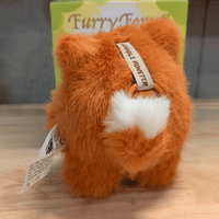 OHKU Giggle Monster plush in fluffy orange creature costume with fluffy white tail and fabric tag visible on wooden table