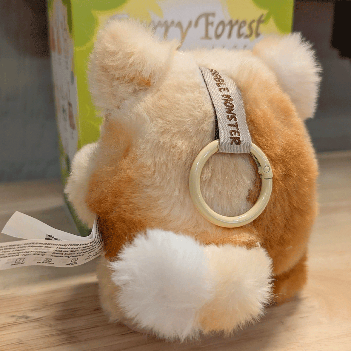 OHKU Giggle Monster plush figure in soft tan and white fur with keychain ring and tag on wooden surface