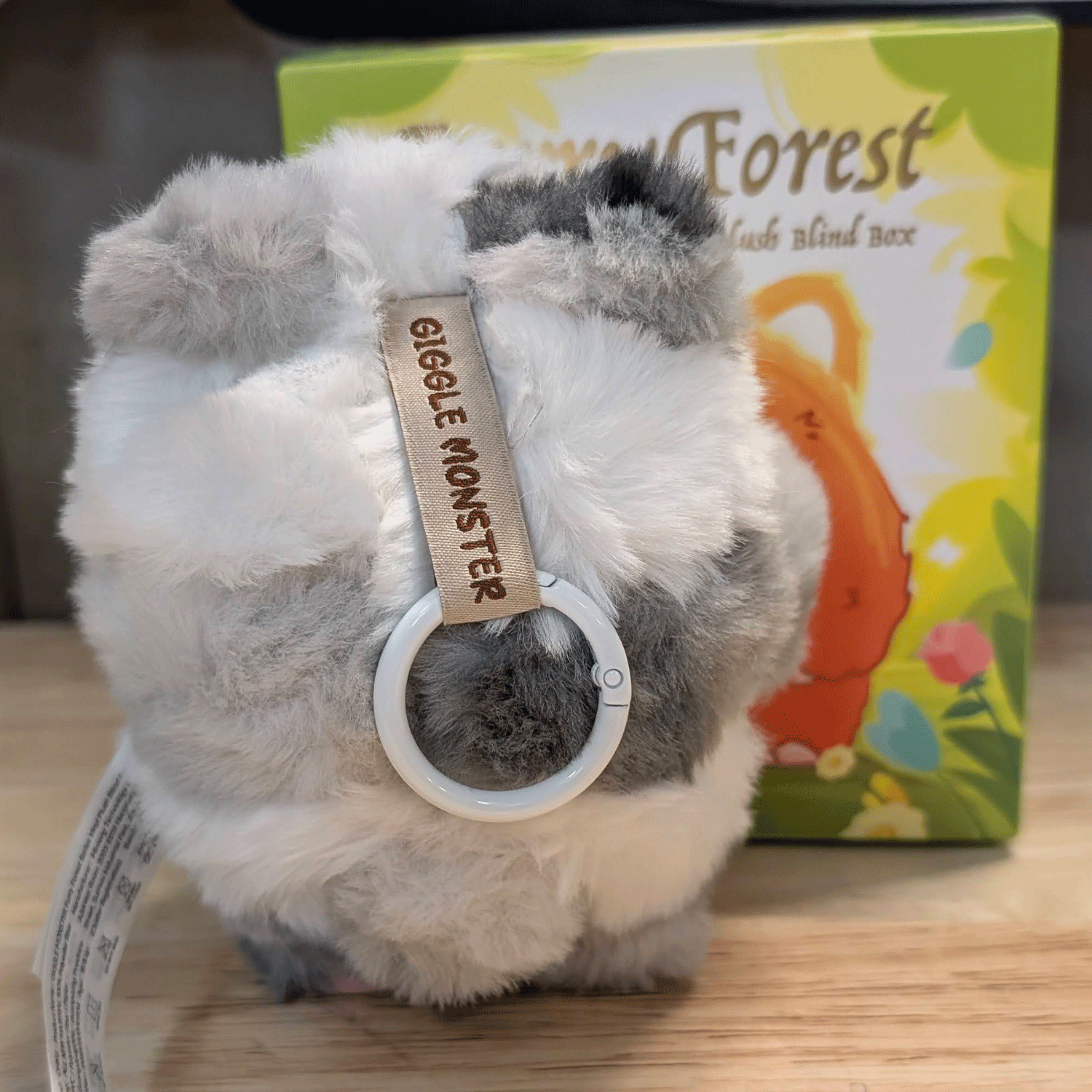 OHKU Giggle Monster plush keychain in soft patchy gray and white fur with Furry Forest blind box packaging behind