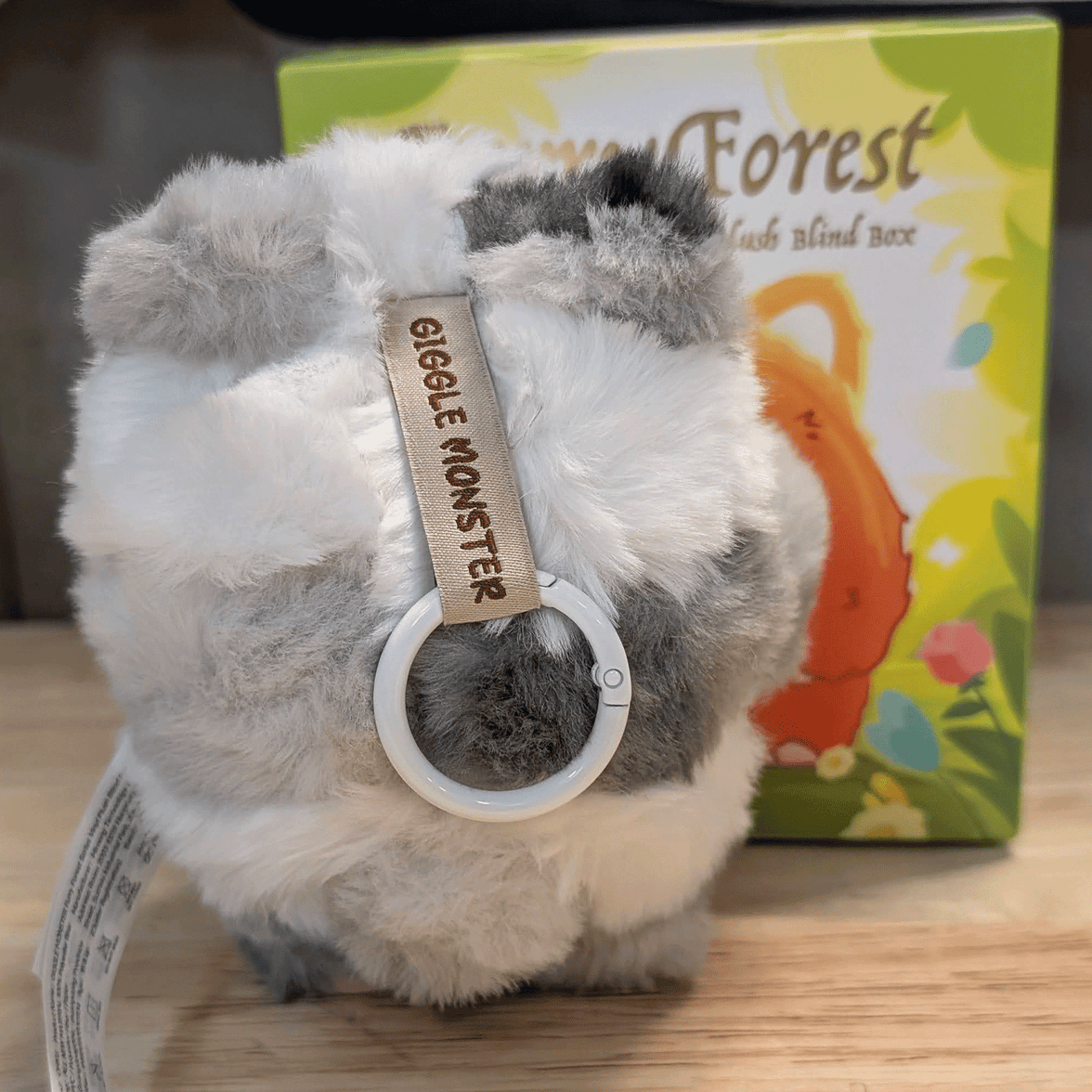 OHKU Giggle Monster plush keychain in soft patchy gray and white fur with Furry Forest blind box packaging behind