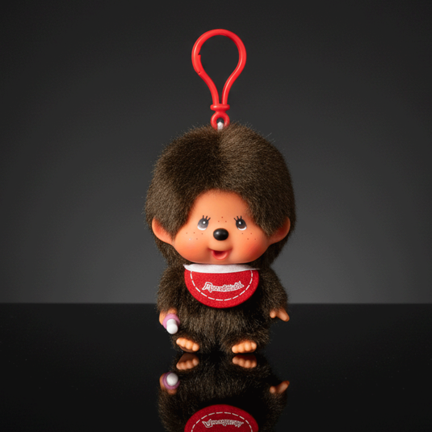 Monchhichi classic boy plush keychain with red bib and thumb in mouth hanging against dark background by Sekiguchi