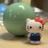 Go Figment Hello Kitty figure with red bow and blue outfit standing next to green capsule on wooden surface