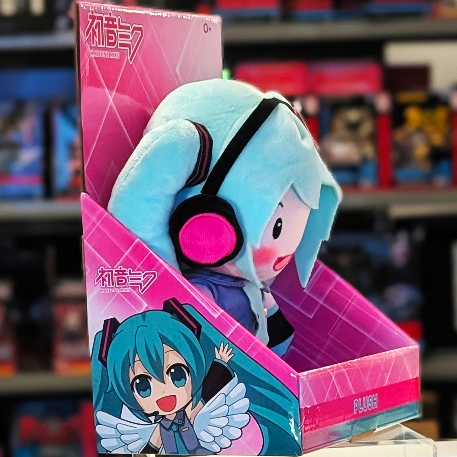 Side view of 12-inch Hatsune Miku plush with teal hair and pink headphones in UCC branded pink display box