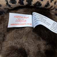 Sekiguchi Monchhichi plush tag close-up showing brand name and materials label on leopard hood and cape stuffed toy