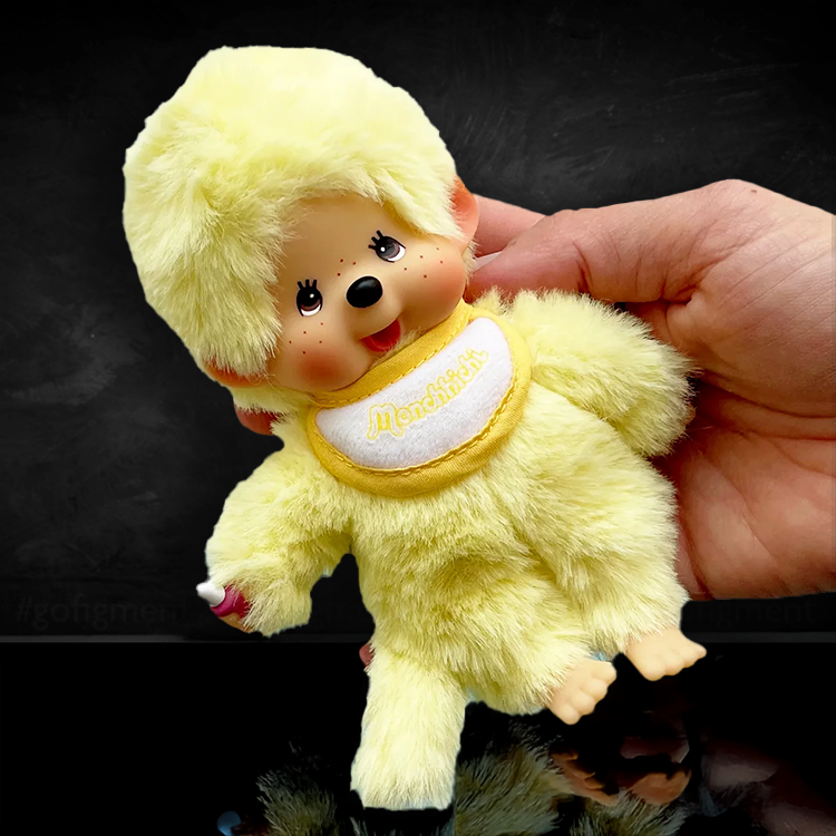 Sekiguchi Monchhichi plush in sunny yellow beanie with soft body and classic face, held by hand on black background