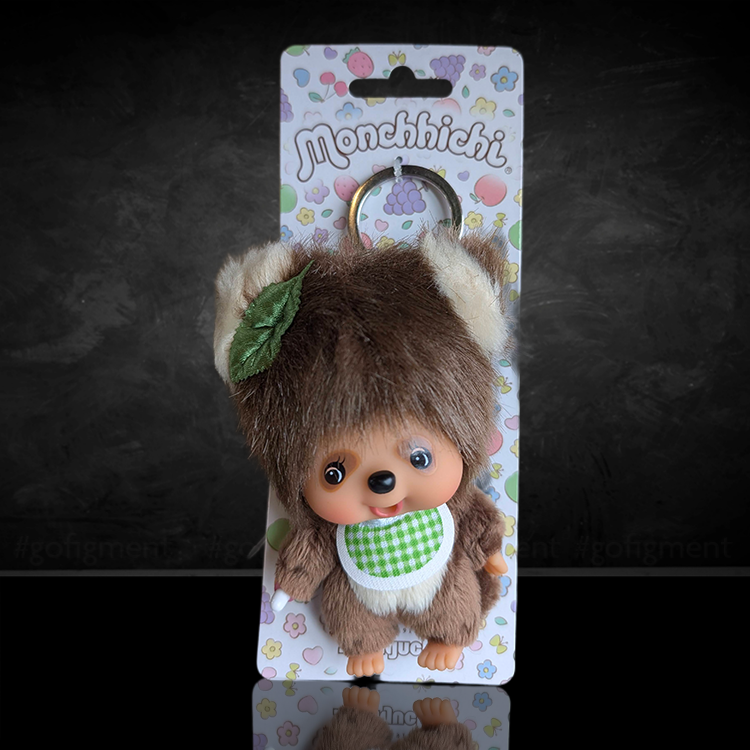 Monchhichi keychain doll with brown fur, green gingham bib, leaf on head, and cute smiling face on colorful card background