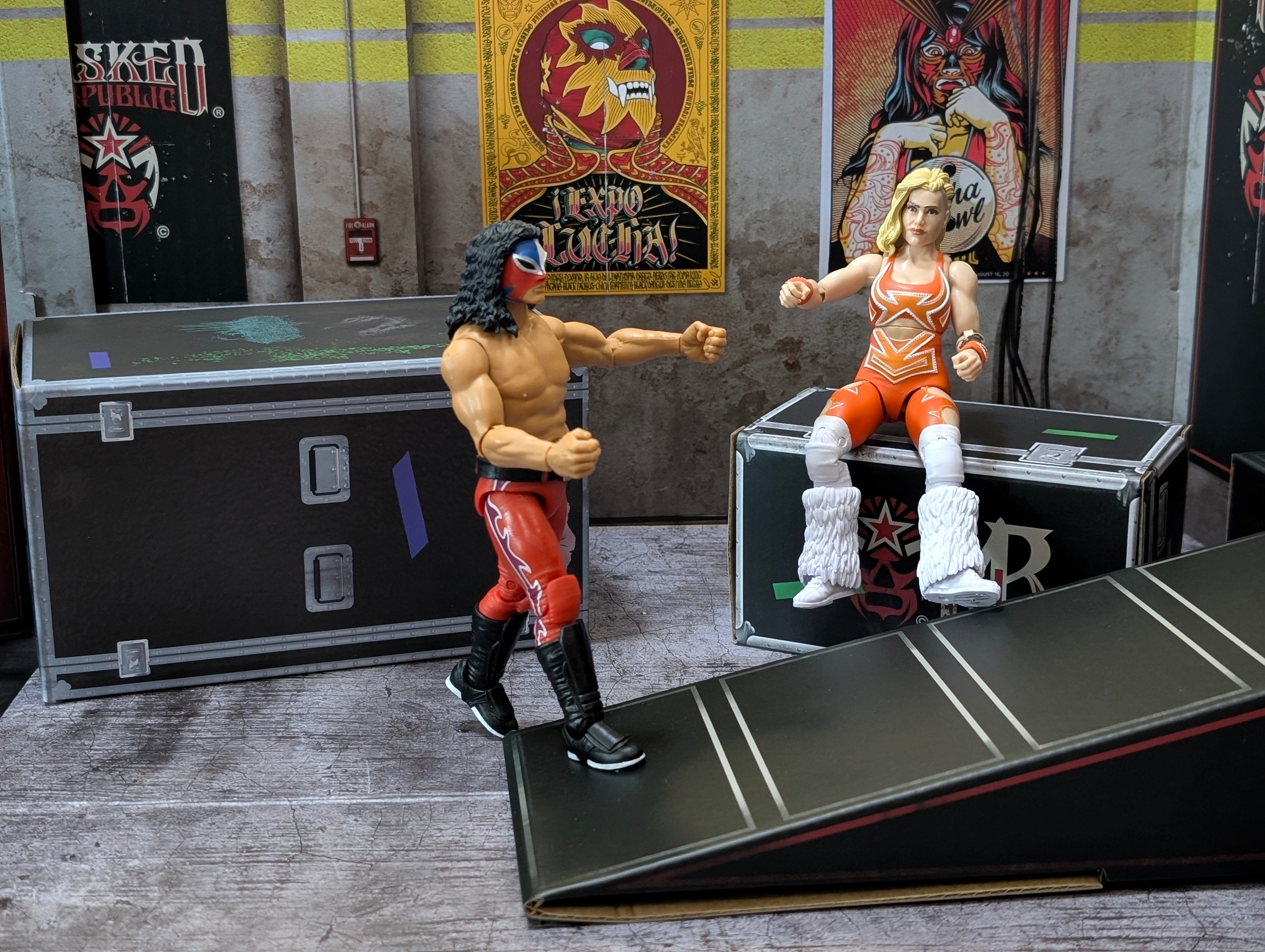 Two wrestling action figures posed near a ramp with event posters and equipment cases in the background