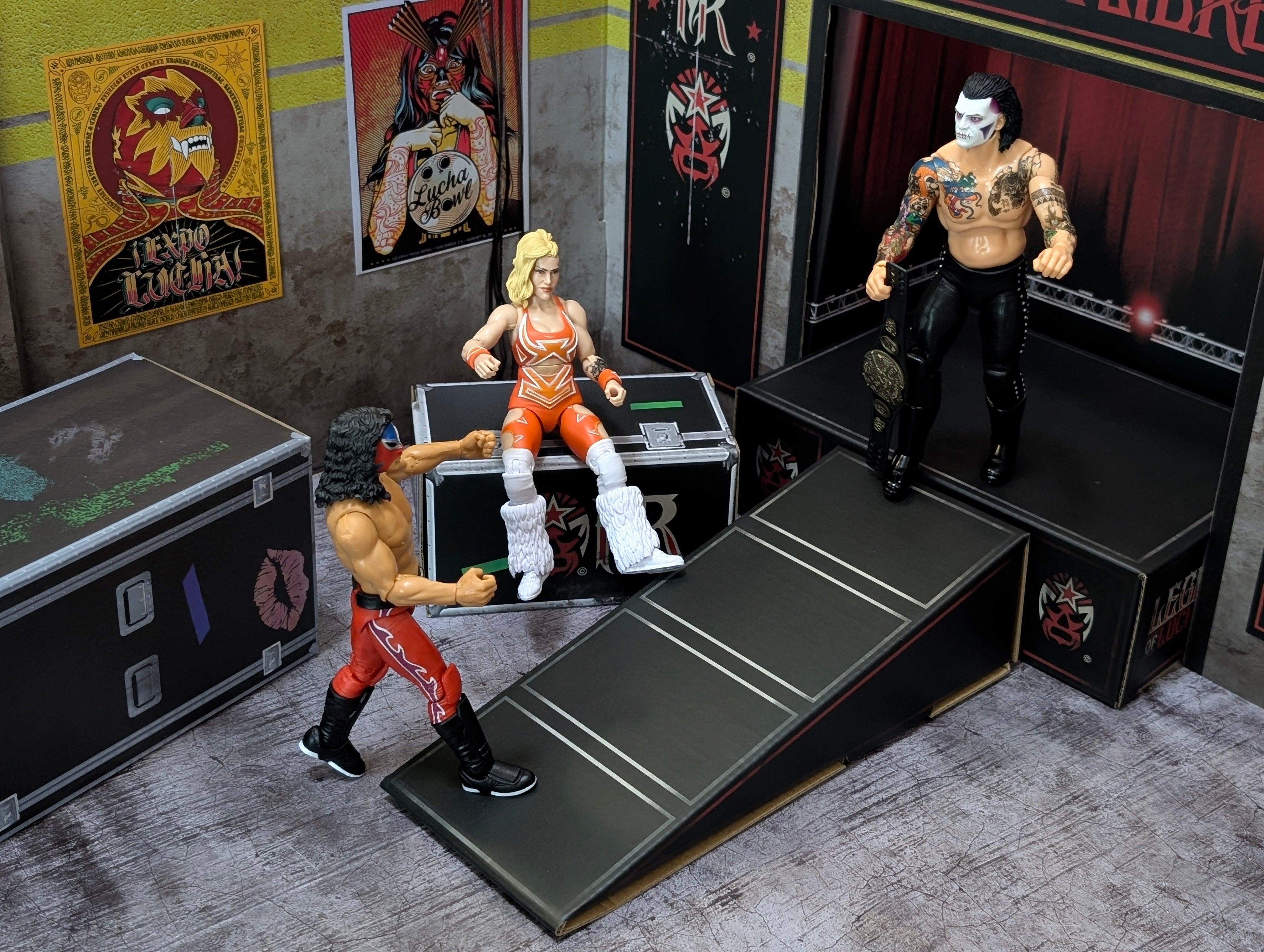 Three wrestling action figures posed near a stage with posters and equipment cases in the background