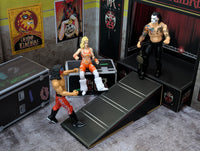 Three wrestling action figures posed near a stage with posters and equipment cases in the background