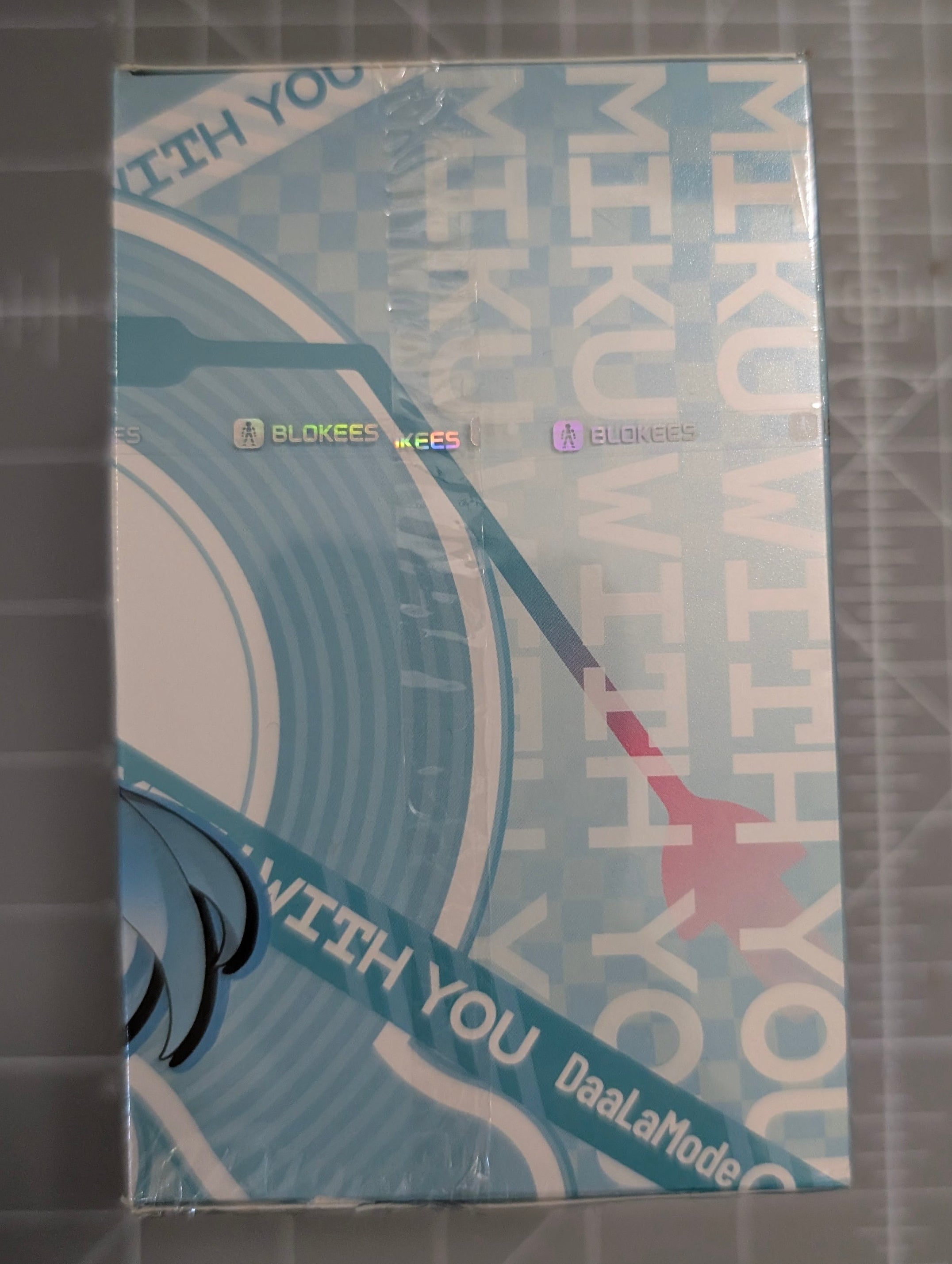 A sealed retail box for a DuaLaMode x BLOOM x TREASURE collaboration, featuring a light blue and white design with a stylized vinyl record and Japanese text, placed on a gray cutting mat.