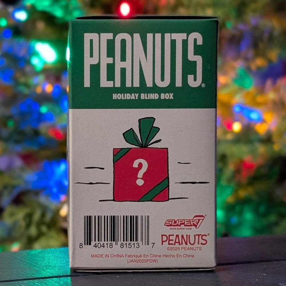 Super7 Peanuts Holiday Blind Box with red gift wrapped in green ribbon and question mark against festive blurred lights background