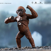 Articulated gorilla figure with textured brown fur and green eyes posed with raised fists on rocky surface against blurred background