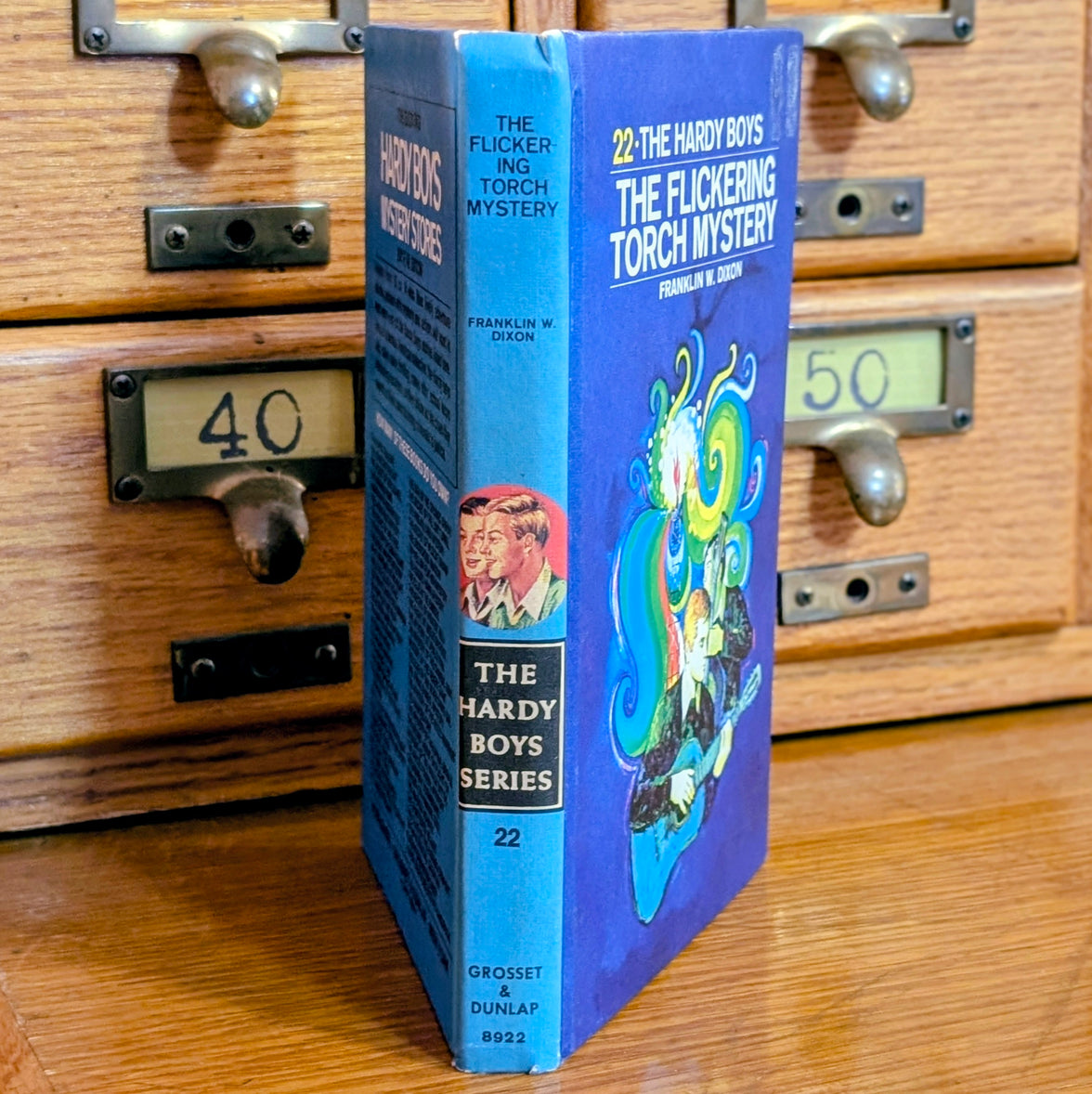 Vintage Hardy Boys book The Flickering Torch Mystery 22 by Grosset & Dunlap with blue back cover standing on wooden drawers