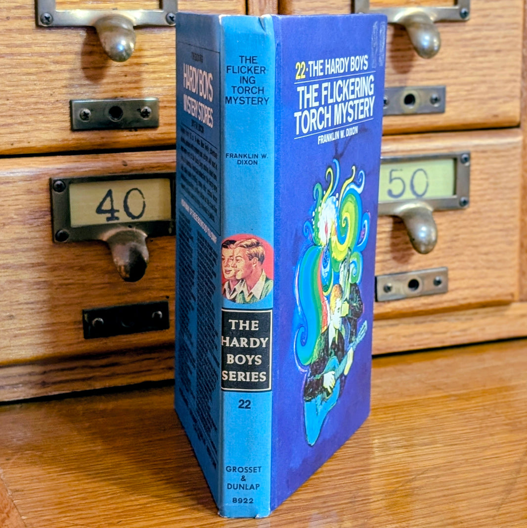 Vintage Hardy Boys book The Flickering Torch Mystery 22 by Grosset & Dunlap with blue back cover standing on wooden drawers